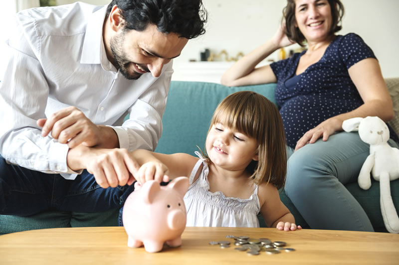 pregnant woman on sofa looking on at man and little girl playing with a piggy bank