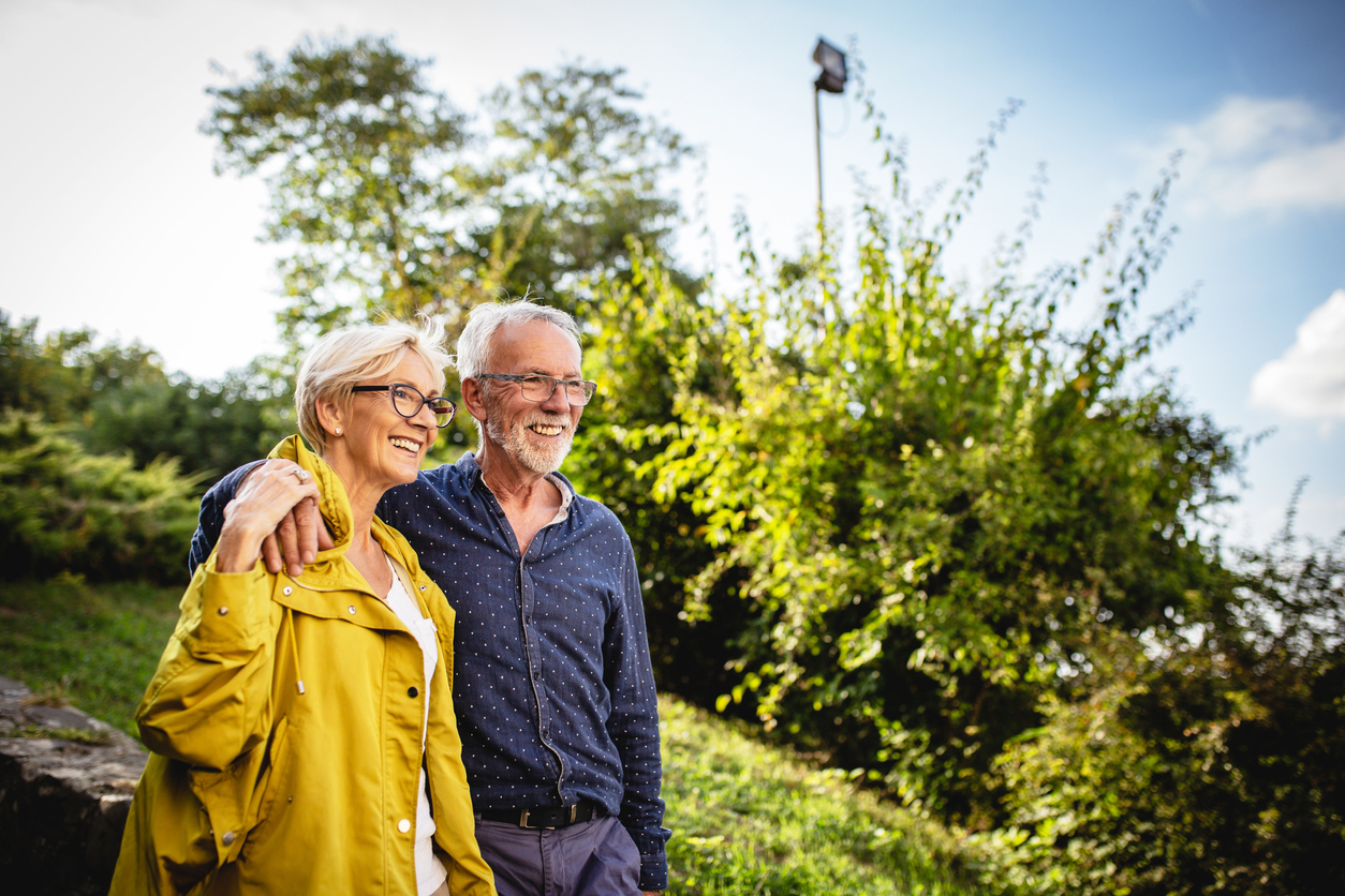 older couple looking out across a view on a sunny day with a green grass and bushes background -Escaping the higher minimum pension age