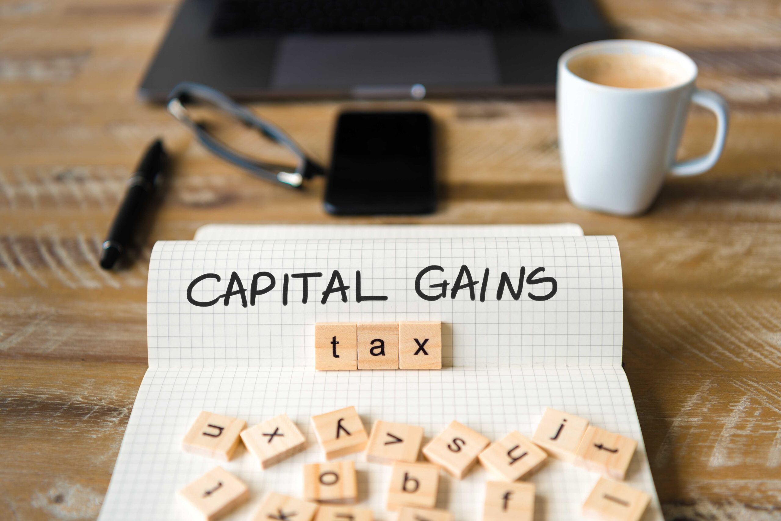 A graphic showing a piece of paper with 'Capital Gains' handwritten on the page and wooden squares with letters on scattered across it. The wooden squares spell out 'tax' beneath 'Capital Gains'.